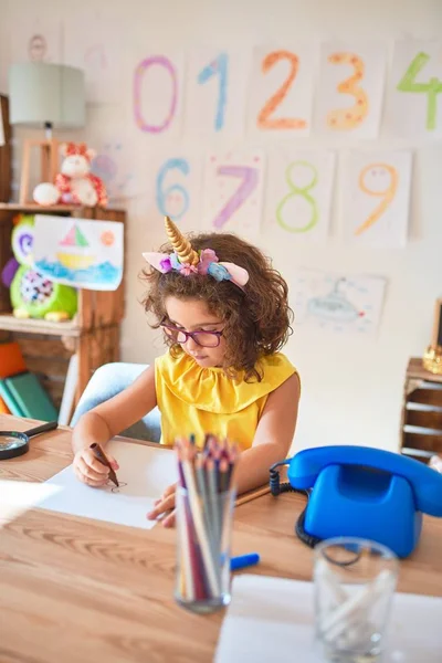 幼儿园时 漂亮的幼儿戴着眼镜 骑着独角兽日记 坐在写字台上 用纸和笔划着记号 — 图库照片