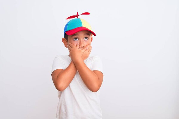 Beautiful kid boy wearing fanny colorful cap with propeller over isolated white background shocked covering mouth with hands for mistake. Secret concept.