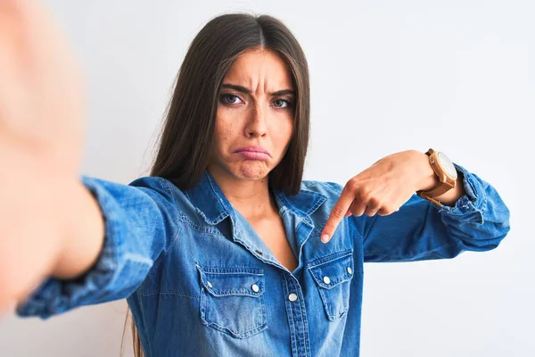 Beautiful woman wearing denim shirt make selfie by camera over isolated white background Pointing down looking sad and upset, indicating direction with fingers, unhappy and depressed.