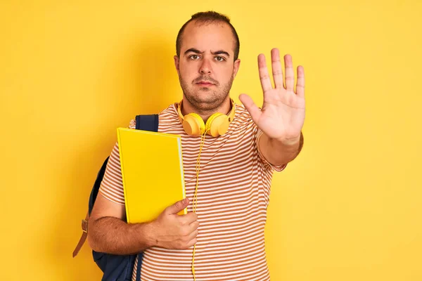 学生们头戴背负式耳机 手持笔记本 背对着孤立的黄色背景 张开双手 做了个句号 表情严肃而自信 做了个防卫手势 — 图库照片