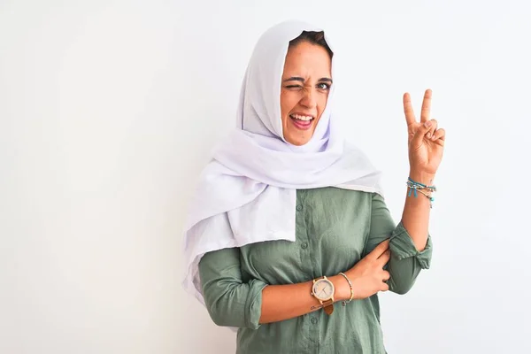 孤立した背景に伝統的なイスラム教徒のヒジャーブを身に着けている若い美しいアラブ人女性は 勝利の兆候を行うカメラで幸せな顔をウィンクで笑顔 第二番 — ストック写真