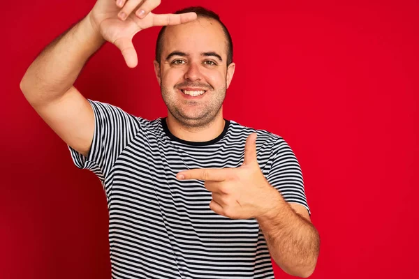 年轻男子穿着条纹海军T恤 站在孤立的红色背景上 微笑着 用双手和手指制作出笑脸的框架 创意和摄影概念 — 图库照片