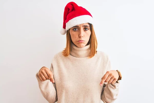 美しい赤毛の女性は 孤立した背景の上にクリスマスの帽子を身に着けている指で方向を示す 悲しいと動揺を下に向けて 不幸なと落ち込んで — ストック写真