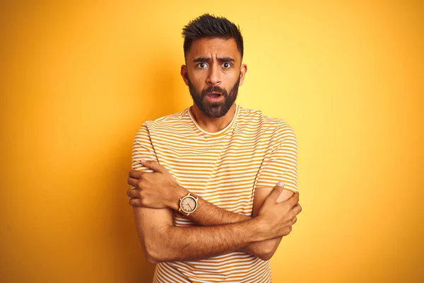 Young indian man wearing t-shirt standing over isolated yellow background shaking and freezing for winter cold with sad and shock expression on face