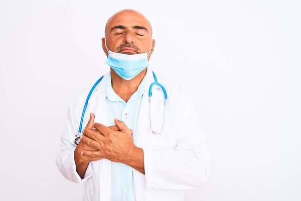閉じられた目と顔に感謝のジェスチャーで胸に手で笑みを浮かべて隔離された白い背景の上に聴診器とマスクを身に着けている中世の医師の男 健康コンセプト — ストック写真