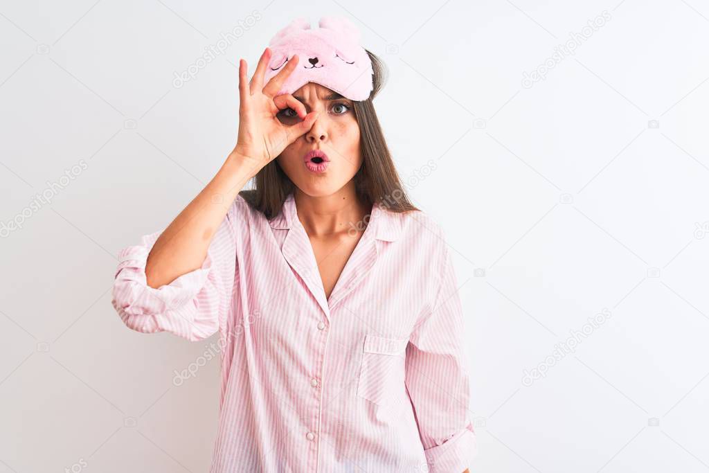 Young beautiful woman wearing sleep mask and pajama over isolated white background doing ok gesture shocked with surprised face, eye looking through fingers. Unbelieving expression.