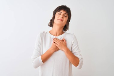 İzole edilmiş beyaz arka plan üzerinde sıradan bir tişört giyen genç ve güzel bir kadın. Elleri göğsünde, gözleri kapalı ve yüzünde minnet dolu bir jest ile gülümsüyor. Sağlık kavramı.