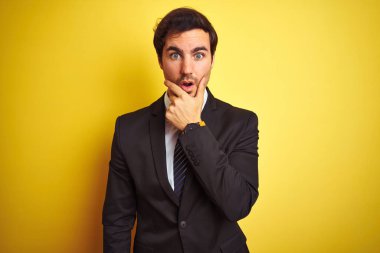 Sarı arka plan üzerinde duran takım elbiseli ve kravatlı genç yakışıklı işadamı elleri çenesinde olan inançsızlıktan, şaşkınlıktan ve şaşkınlıktan büyülenmiş görünüyor.