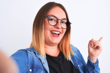 Kot gömlek ve gözlük takan güzel bir kadın izole edilmiş beyaz arka plan üzerinde selfie yapar. El ve parmak kenarı işaret ederek çok mutlu olur.
