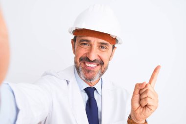 Miğfer takan orta yaşlı mühendis, izole edilmiş beyaz arka plan üzerinde selfie çeker. Elini ve parmağını kenarı göstererek mutlu olur.