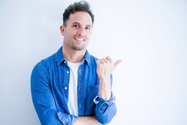 İzole edilmiş beyaz arka plan üzerinde duran kot gömlek giyen genç yakışıklı adam mutlu bir yüz ifadesiyle gülümsüyor ve başparmağıyla yan tarafı işaret ediyor..