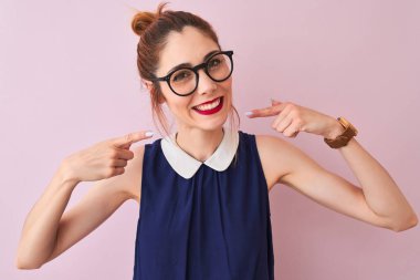 Kırmızı saçlı kadın zarif bir elbise ve gözlük takıyor pembe arka planda gülümsüyor ve parmakları ve ağzıyla işaret ediyor. Diş sağlığı konsepti.