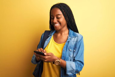 Zeki telefon kullanan Afrikalı Amerikalı bir bayan mutlu bir yüzü olan, kendine güvenen bir gülümseme ile izole edilmiş sarı arka planda duruyor ve dişlerini gösteriyor.