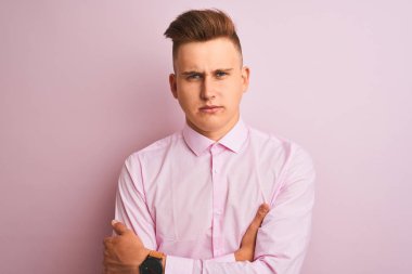 Soyutlanmış pembe arka plan üzerinde duran zarif gömlekli genç yakışıklı işadamı şüpheci ve gergin, yüzünde çapraz kollu tasvip etmeyen bir ifade. Negatif kişi.