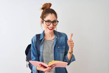 Sırt çantası takan kızıl saçlı kız, izole edilmiş beyaz arka plan üzerine kitap okuyor. Gülümseyerek mutlu. Tamam işareti yap, parmaklarınla başparmağını kaldır.