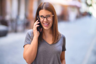 Mutlu ve kendinden emin gülümseyen genç ve güzel bir kadın. Yüzünde gülümsemeyle kasaba caddesinde akıllı telefonun başında konuşmak.