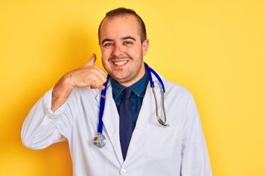 Paltolu ve steteskoplu genç bir doktor izole edilmiş sarı arka planda durup telefonla el ve parmaklarıyla telefonla konuşuyor gibi gülümsüyor. İletişim kavramları.