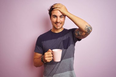 Dövmeli ve kahve içen genç yakışıklı bir adam izole edilmiş pembe arka planda, elleri başının üstünde, utanç ve şaşkınlık dolu, sinirli ve hayal kırıklığına uğramış bir halde. Hata için korku ve üzüntü.