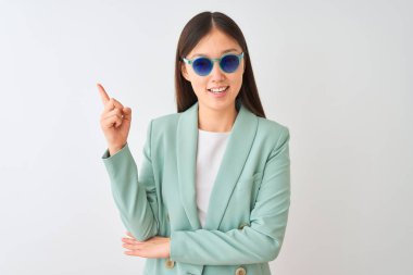 İzole edilmiş beyaz arka planda ceket ve güneş gözlüğü takan Çinli iş kadını elini ve parmağını kenarı işaret ederek çok mutlu.