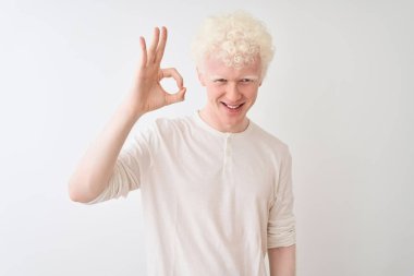 İzole edilmiş beyaz arka plan üzerinde sıradan tişörtler giyen genç albino sarışın adam el ve parmaklarıyla tamam işareti yaparken gülümsüyor. Başarılı ifade.