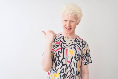 Genç, albino, renkli tişört giyen, izole edilmiş beyaz arka planda duran mutlu yüzüyle gülümseyen ve başparmağıyla yan tarafı gösteren sarışın bir adam..