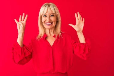 Orta yaşlı bir kadın zarif bir gömlek giyiyor. Kırmızı arka plan üzerinde duruyor. Dokuz numarayı gösteriyor ve gülümsüyor..