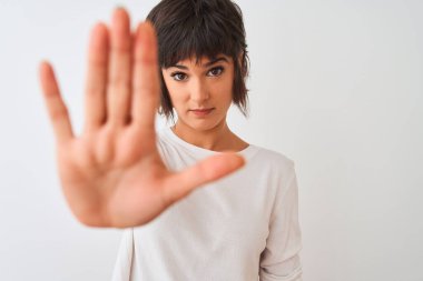 İzole edilmiş beyaz arka plan üzerinde duran, ciddi ve kendinden emin ifadeyle dur işareti yapan, sıradan bir tişört giyen genç ve güzel bir kadın.