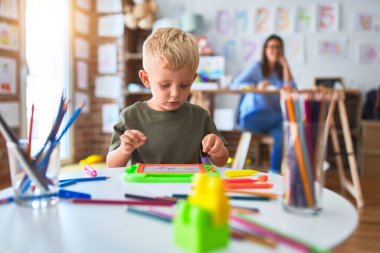 Genç beyaz çocuk öğretmeniyle oyun okulunda oynuyor. Anne ve oğlu oyun odasında renkli kalemlerle çizim yapıyor. Arka planda oturan genç kadın da masada oturuyor..