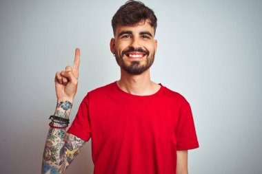 İzole edilmiş beyaz arka plan üzerinde duran kırmızı tişörtlü genç adam bir numaralı parmağını gösterirken kendine güvenen ve mutlu bir şekilde gülümsüyor..