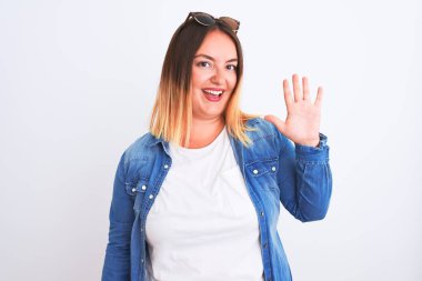 İzole edilmiş beyaz arka plan üzerinde duran kot gömlek giyen güzel bir kadın beş numaralı parmaklarıyla işaret ederken kendinden emin ve mutlu bir şekilde gülümsüyor..