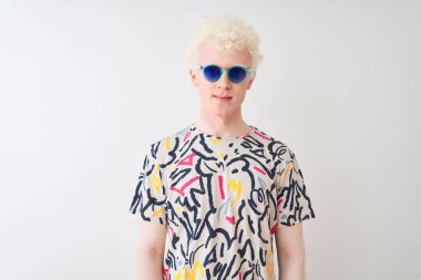 Genç albino sarışın adam yüzünde ciddi ifade ile izole kırmızı arka plan üzerinde renkli t-shirt ve güneş gözlüğü giyiyor. Kameraya basit ve doğal bir bakış.