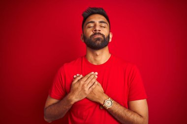 İzole edilmiş kırmızı arka plan üzerinde tişört giyen genç, yakışıklı Hintli adam elleri göğsünde, gözleri kapalı ve yüzünde minnet dolu bir jest ile gülümsüyor. Sağlık kavramı.