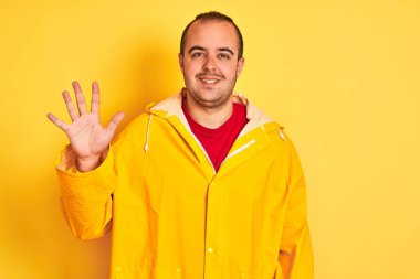 Sarı arka plan üzerinde duran yağmurluk giyen genç adam beş numaralı parmaklarıyla gülümseyerek kendinden emin ve mutlu bir şekilde işaret ediyor..