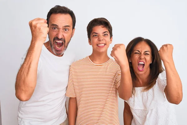 Family of three, mother, father and son standing over white isolated background angry and mad raising fist frustrated and furious while shouting with anger. Rage and aggressive concept.