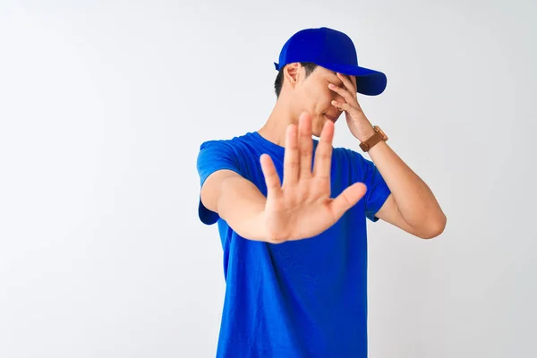 Chinese deliveryman wearing blue t-shirt and cap standing over isolated white background covering eyes with hands and doing stop gesture with sad and fear expression. Embarrassed and negative concept.