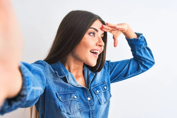 Beautiful woman wearing denim shirt make selfie by camera over isolated white background very happy and smiling looking far away with hand over head. Searching concept.