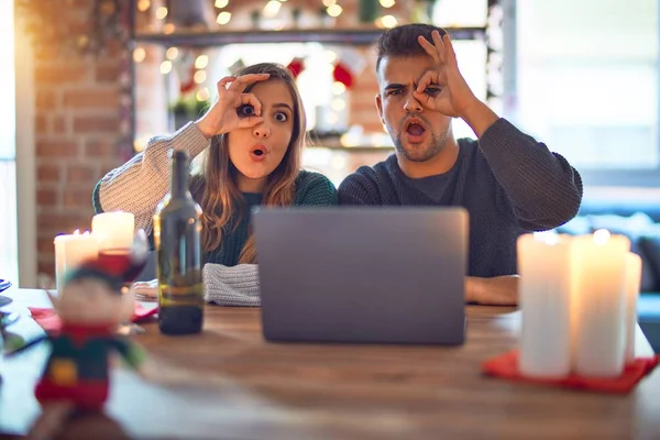 Young beautiful couple sitting using laptop around christmas decoration at home doing ok gesture shocked with surprised face, eye looking through fingers. Unbelieving expression.