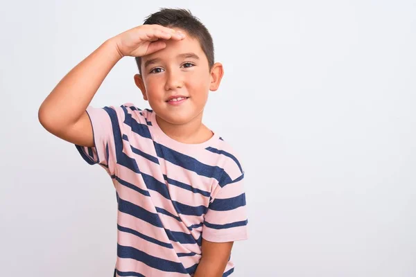 Beautiful kid boy wearing casual striped t-shirt standing over isolated white background very happy and smiling looking far away with hand over head. Searching concept.