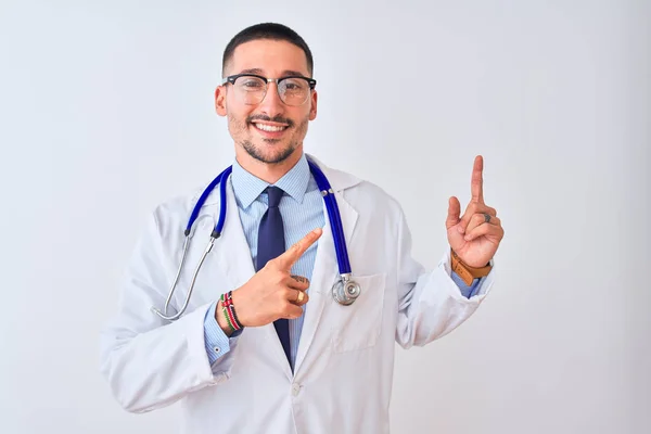 年轻的医生 戴着听诊器 背对着孤立的背景微笑着 看着摄像机 用双手和手指指向旁边 — 图库照片