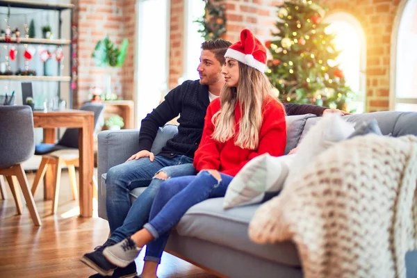 年轻夫妇头戴圣诞月桂冠 坐在沙发上 坐在圣诞树旁 面带微笑 悠闲自在地摆姿势 — 图库照片