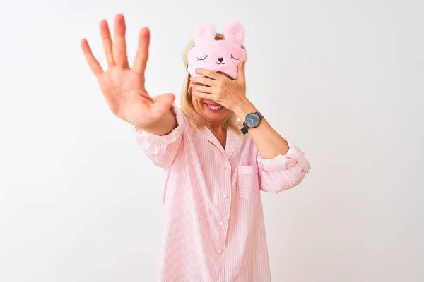 Middle age woman wearing sleep mask and pajama over isolated white background covering eyes with hands and doing stop gesture with sad and fear expression. Embarrassed and negative concept.