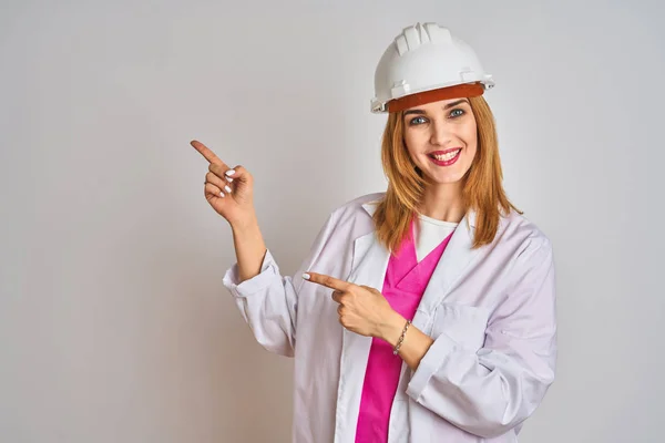 红头发的高加索女工程师头戴安全帽 面带微笑地看着摄像机 用双手和手指指向两侧 — 图库照片