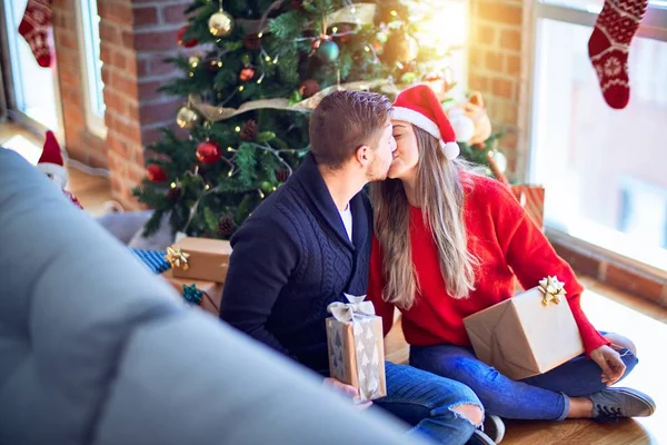 年轻漂亮的夫妇面带微笑 既快乐又自信 坐在地板上拿着礼物 在家里的圣诞树旁拥抱和亲吻 — 图库照片