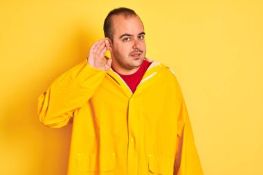 İzole edilmiş sarı arka planda dikilen yağmurluk giymiş genç bir adam kulağına fısıldayarak dedikoduları ve dedikoduları dinliyor. Sağırlık kavramı.