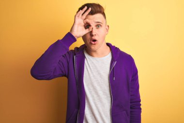 Mor eşofman üstü giyen yakışıklı genç adam izole edilmiş sarı arka planda dikiliyor. Şaşırmış yüz ifadesiyle, parmaklarından bakan gözleriyle şok oluyor. İnançsız ifade.