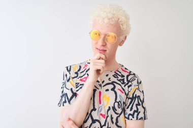 Renkli tişört ve güneş gözlüğü takan genç albino bir adam izole edilmiş kırmızı arka planda, elleri çenesinde soru sorarken, düşünceli bir ifade. Düşünceli bir yüzle gülümsüyor. Şüphe kavramı.