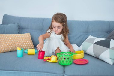 Sevimli sarışın bebek kanepede oturuyor evde yemek ve çatal bıçakla yemek pişiriyor.