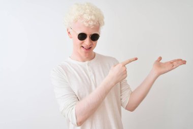 İzole edilmiş beyaz arka planda, tişört ve güneş gözlüğü takan genç albino adam kameraya hayret ediyor ve gülümsüyor..