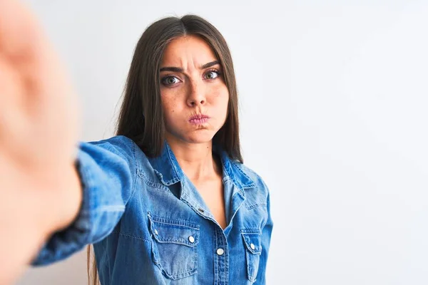 Beautiful woman wearing denim shirt make selfie by camera over isolated white background puffing cheeks with funny face. Mouth inflated with air, crazy expression.