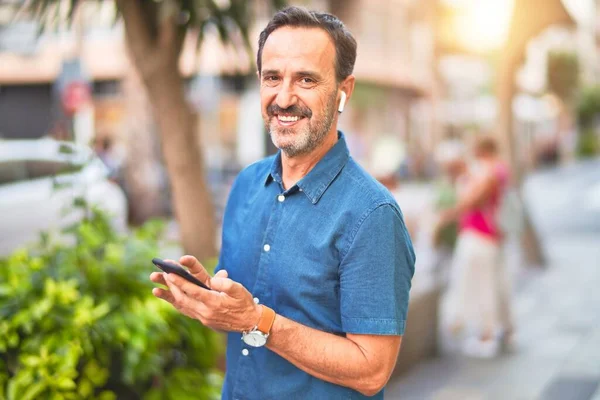 中年英俊商人站在街上 用智能手机和耳机笑着 — 图库照片